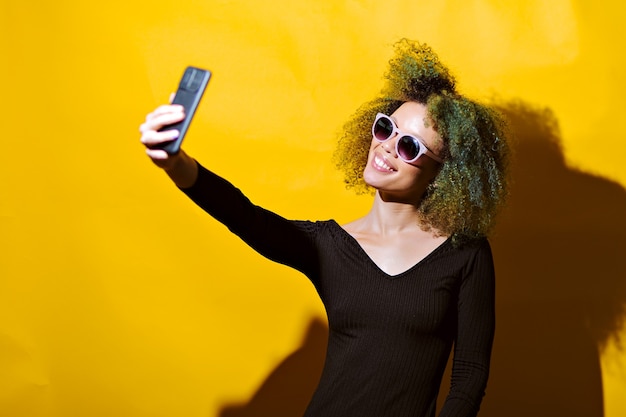 Afro kobieta robi selfie w okularach przeciwsłonecznych na żółtym tle