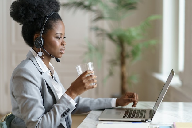 Afro Pracownik Kobieta Patrząc Na Ekran Laptopa, Pracujący W Domowym Biurze, Woda Pitna Ze Szklanki.