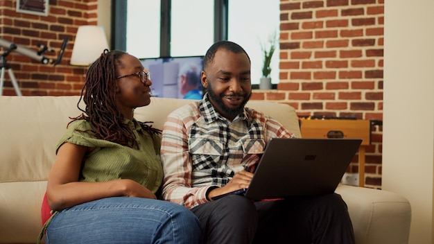 Afroamerykanie oglądają filmy online na laptopie, relaksują się razem na kanapie i dobrze się bawią. Przeglądanie internetu na komputerze i cieszenie się wolnym czasem, śmiejąc się w domu.