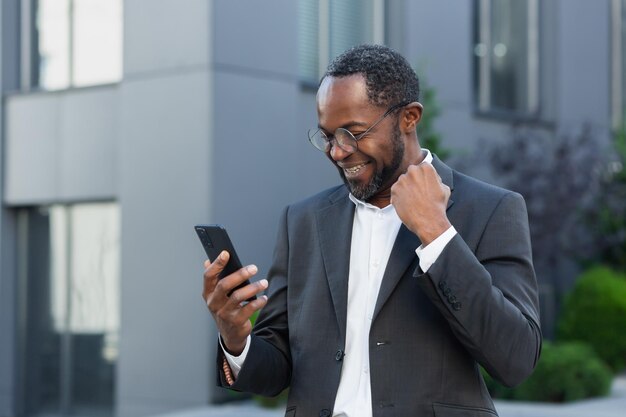 Afroamerykanin biznesmen szef poza nowoczesnym biurowcem za pomocą telefonu starszy mężczyzna świętuje