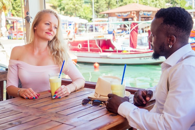 Afroamerykanin i kobieta patrzą z miłością na siebie w letniej tropikalnej kawiarni i piją świeży pomarańczowy koktajl