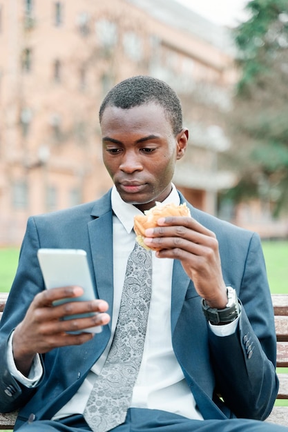 Afroamerykanin je burgera w parku na świeżym powietrzu podczas korzystania ze swojego smartfona