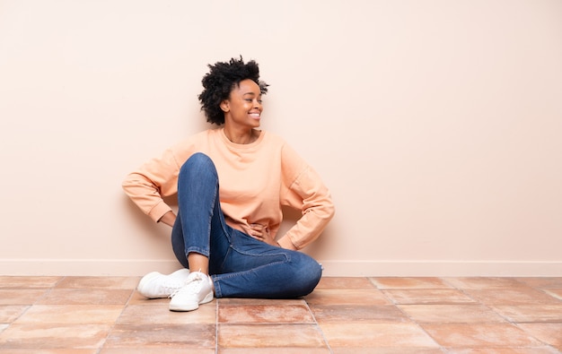Afroamerykanin kobieta siedzi na podłodze, pozowanie z rękami na biodrze i uśmiechnięty