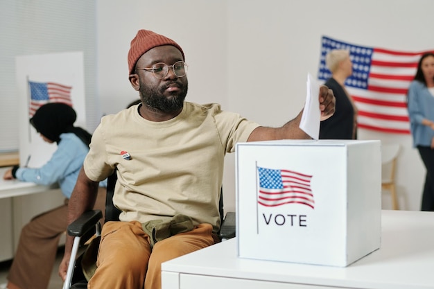 Afroamerykanin młody człowiek z niepełnosprawnością wkłada kartę do głosowania do pudełka