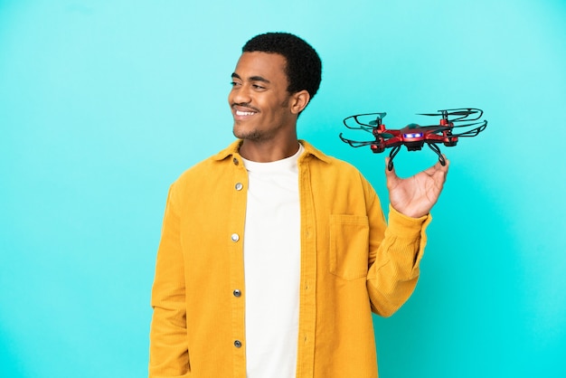 Afroamerykanin, przystojny mężczyzna, trzymający drona na odosobnionym niebieskim tle, patrząc w bok i uśmiechnięty