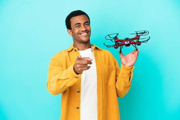 Afroamerykanin, przystojny mężczyzna, trzymający drona na odosobnionym niebieskim tle, wskazujący przód ze szczęśliwym wyrazem twarzy