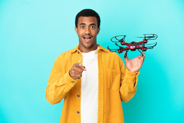Afroamerykanin przystojny mężczyzna trzymający drona na odosobnionym niebieskim tle zaskoczony i wskazujący przód