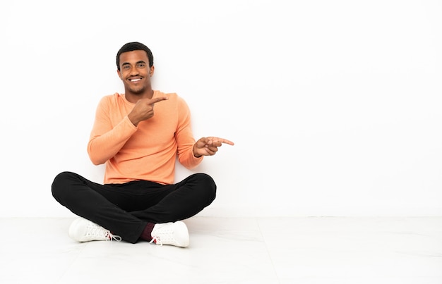 Afroamerykanin siedzący na podłodze na białym tle copyspace zaskoczony i wskazujący bok