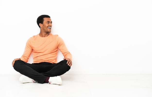 Afroamerykanin siedzący na podłodze nad odosobnioną ścianą copyspace, śmiejący się w pozycji bocznej