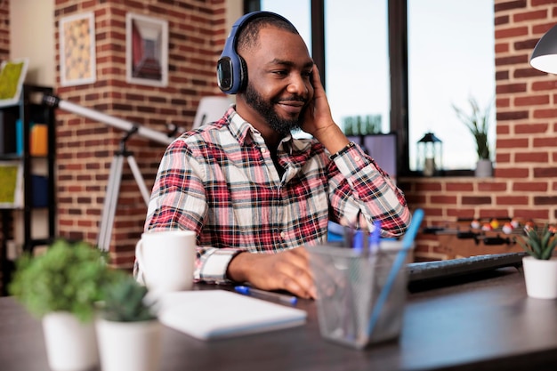 Afroamerykanin słucha podcastu lub muzyki na słuchawkach, korzystając z pokazu online w mediach społecznościowych na zestawie słuchawkowym, podczas gdy on pracuje nad start-upem z domu. Wykonywanie zdalnej pracy na komputerze.