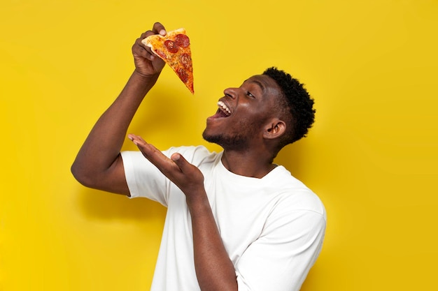 Afroamerykanin w białej koszulce trzyma kawałek pizzy z otwartymi ustami na żółtym tle