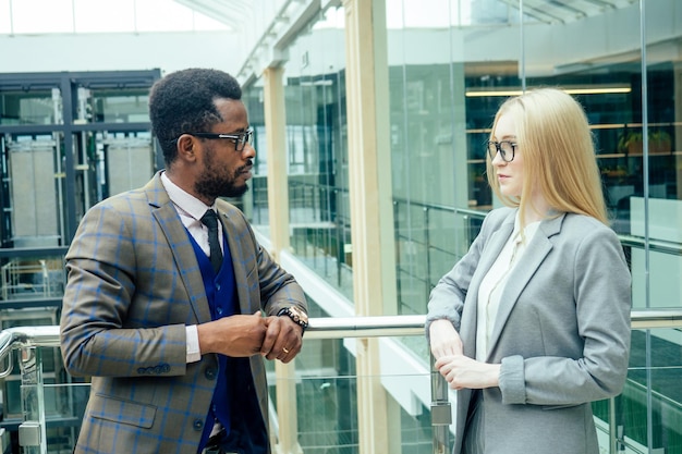 Afroamerykanin w białej koszuli w brązowej kurtce pozuje rozmawia z biznesową blondynką z kaukazu w szarym garniturze w nowoczesnym biurze z przeszklonymi oknami w centrum handlowym