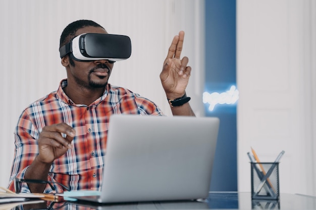 Afroamerykanin w okularach VR, pracujący na laptopie w wirtualnej rzeczywistości, siedzący przy biurku