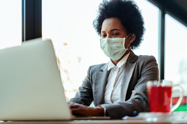 Afroamerykanka bizneswoman z maską ochronną na twarzy pracuje na laptopie siedząc w kawiarni