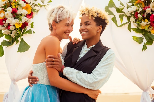 Zdjęcie afroamerykanka pan młody w czarnym garniturze i szczęśliwa kaukaska blond panna młoda kochająca się razem na ceremonii na plaży pod kwiatowym łukiem