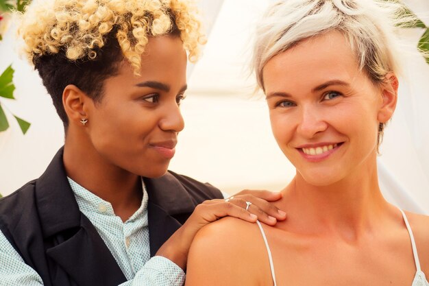 Afroamerykanka pan młody w czarnym garniturze i szczęśliwa kaukaska blond panna młoda kochająca się razem na ceremonii na plaży pod kwiatowym łukiem