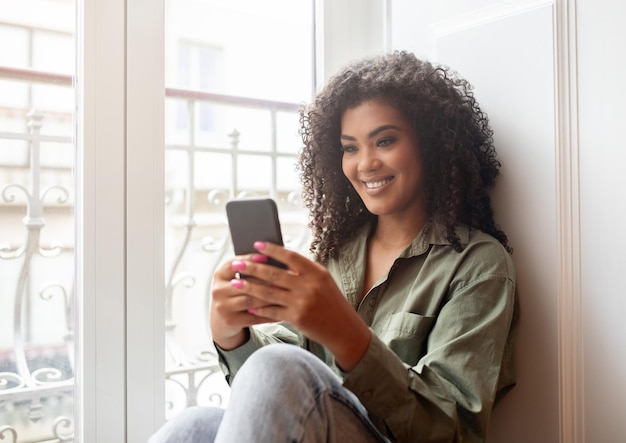 Afroamerykanka pisze SMS-y na smartfonie siedzącym na parapecie w pomieszczeniu