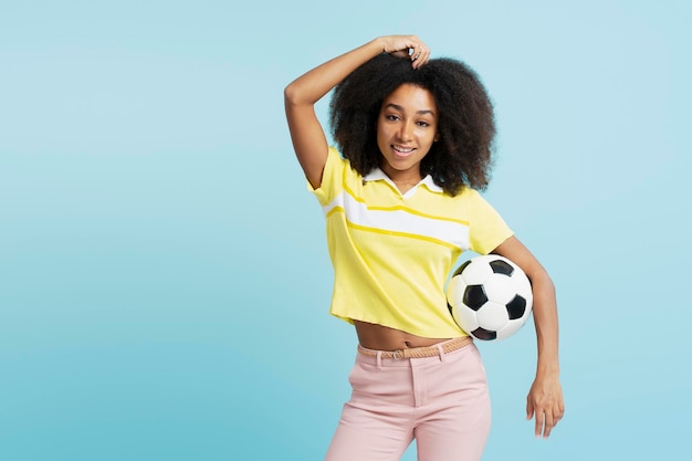 Afroamerykanka ubrana w swobodny strój, trzymająca piłkę nożną i patrząca na kamerę pozowanie na białym tle