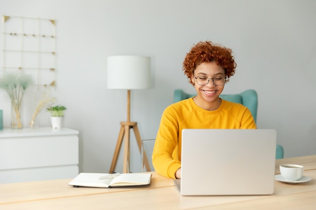 Afroamerykanka używająca laptopa w domowym biurze patrząca na ekran, pisząca na czacie, czytająca, pisząca e-mail