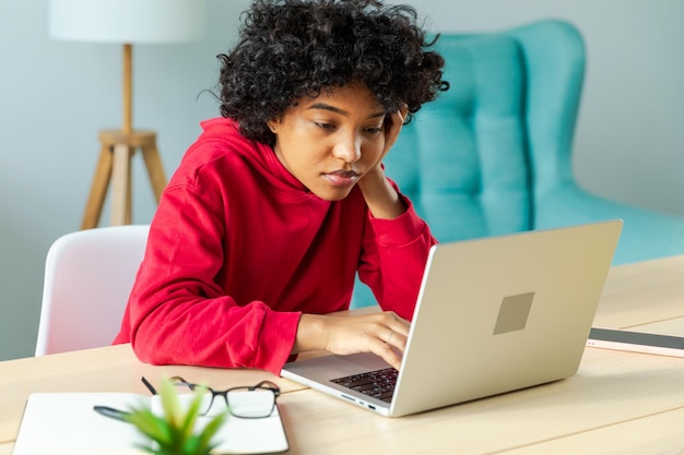 Afroamerykanka używająca laptopa w domowym biurze, patrząca na ekran, pisząca na czacie, czytająca, pisząca