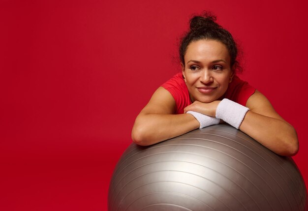 Afroamerykanka wysportowana ładna kobieta relaksuje się na piłce fitness po treningu na białym tle na czerwonym tle z miejscem na kopię