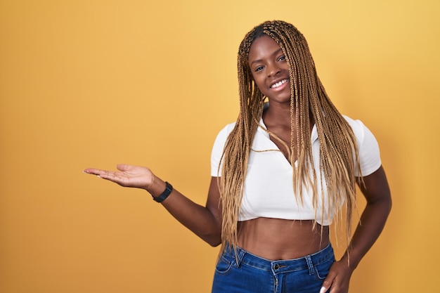 Afroamerykanka z plecionymi włosami, stojąca na żółtym tle, uśmiechnięta radośnie prezentująca się i wskazująca dłonią patrząc w kamerę
