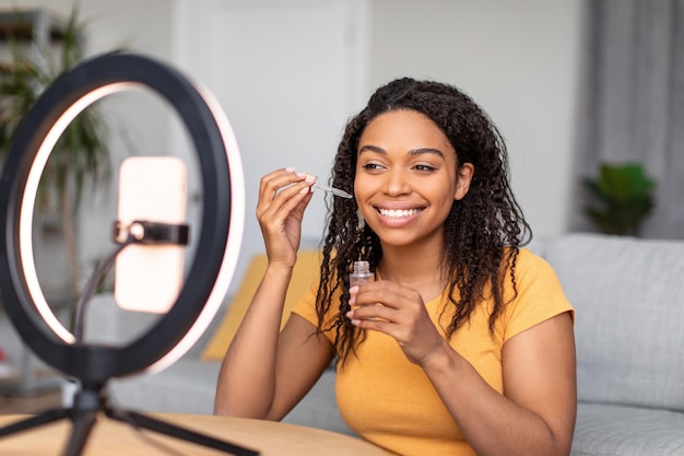 Afroamerykańska blogerka kosmetyczna tworząca wideo na telefonie pokazujące serum do twarzy i reklamy
