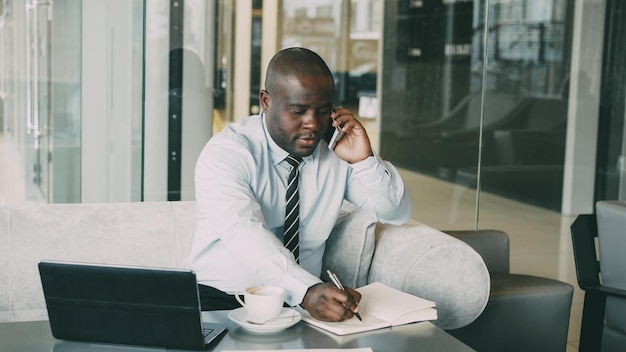 Afroamerykański biznesmen rozmawia na smartfonie i odkłada informacje biznesowe w swoim notatniku, siedząc w nowoczesnej kawiarni z laptopem i kawą na stole