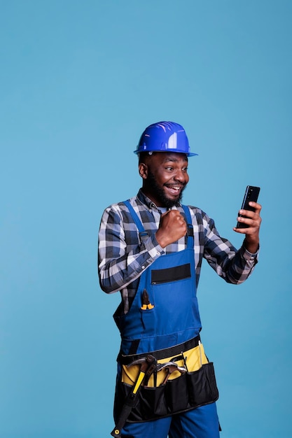 Afroamerykański budowniczy podekscytowany wiadomością o nowym kontrakcie budowlanym otrzymanym przez rozmowę wideo na telefon komórkowy. Wykonawca świętuje z emocji trzymając urządzenie mobilne na niebieskim tle.
