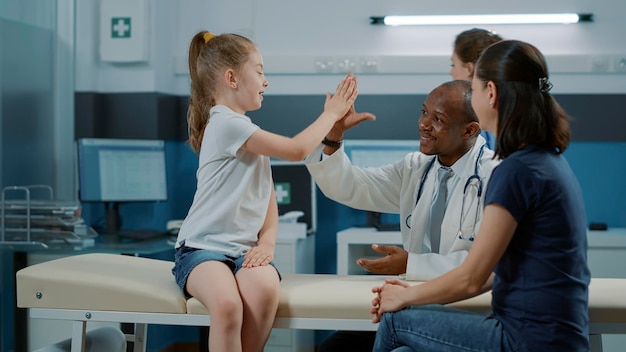 Afroamerykański medyk przybija piątkę małemu dziecku po pomyślnym badaniu kontrolnym. Wesoły dzieciak i lekarz doceniają pracę zespołową na wizytę konsultacyjną z matką w biurze.