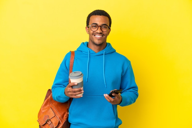 Afroamerykański student mężczyzna nad odosobnioną żółtą ścianą trzymający kawę na wynos i telefon komórkowy