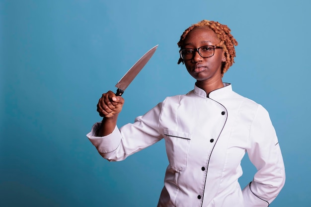 Afrykańska amerykańska szefowa kuchni z kręconymi włosami w mundurze pozuje z ostrym nożem przed kamerą. Profesjonalny kucharz pokazujący naczynie kuchenne w studio nakręcony na niebieskim tle.