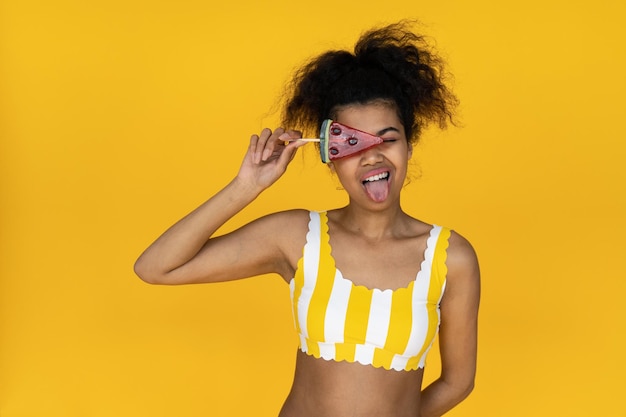 Afrykańska dziewczyna pokazująca język trzymający cukierki na żółtym tle letniego studia