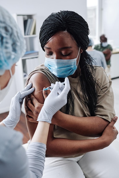 Zdjęcie afrykańska dziewczyna w masce ochronnej dostaje szczepionkę w ramię podczas siedzenia w szpitalu podczas pandemii