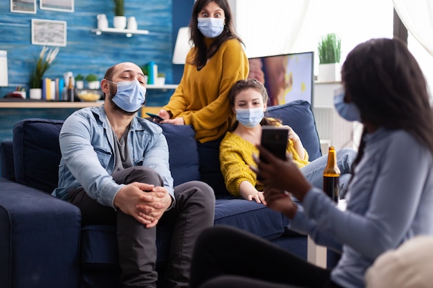 Afrykańska kobieta pokazuje śmieszny klips na smartfonie wychodzę w domu w salonie nosząc maskę zapobiegającą koronawirusowi podczas globalnej pandemii utrzymujący dystans społeczny.