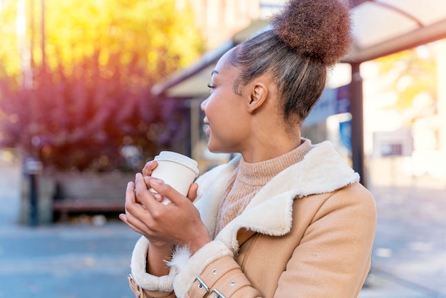 Afrykańska kobieta trzyma filiżankę kawy Uśmiechnięta brunetka z kręconymi włosami w brązowym swetrze czeka na tramwaj