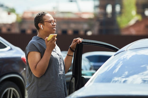 Afrykańska kobieta w bezprzewodowych słuchawkach rozmawia przez telefon komórkowy, stojąc na parkingu na zewnątrz
