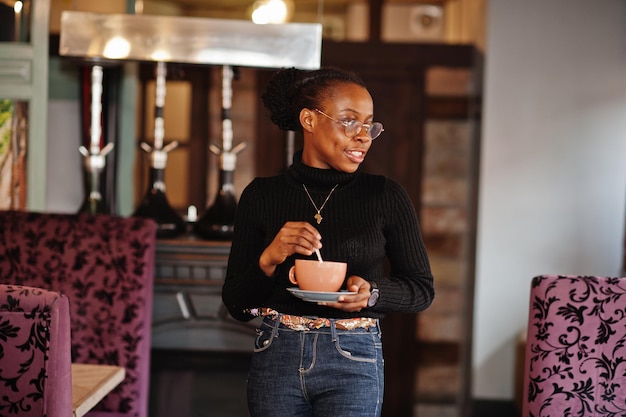 Afrykańska kobieta w czarnym swetrze i okularach pozowała w kawiarni z kubkiem gorącego napoju