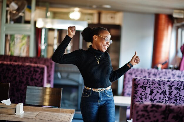 Afrykańska kobieta w czarnym swetrze pozowała w kawiarni i pokazuje ręką kciuk.