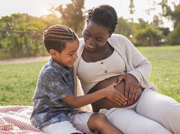 Zdjęcie afrykańska matka w ciąży i syn rasy mieszanej dotykają brzucha w parku miejskim - rodzinna miłość