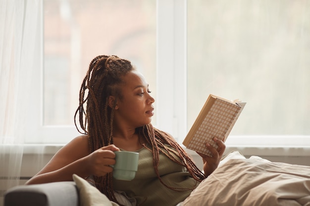Afrykańska młoda kobieta leży na kanapie, pijąc kawę i czytając ciekawą książkę w domu