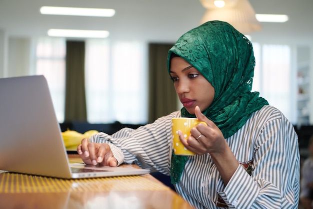afrykańska muzułmańska biznesowa kobieta ubrana w zielony hidżab pijący herbatę podczas pracy na komputerze przenośnym w strefie relaksu w nowoczesnym biurze uruchamiania na otwartym planie. Różnorodność, koncepcja wielorasowa