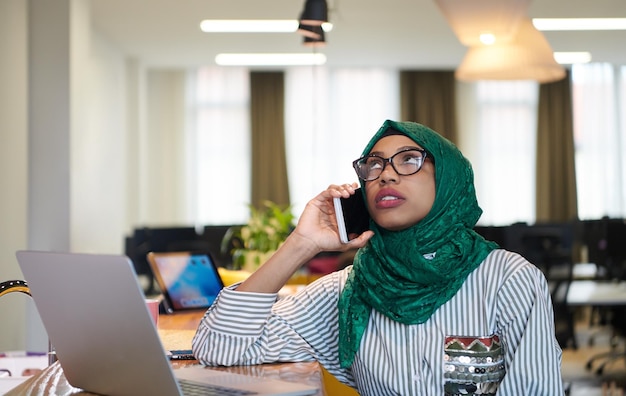afrykańska muzułmańska biznesowa kobieta ubrana w zielony hidżab przy użyciu telefonu komórkowego podczas pracy na komputerze przenośnym w strefie relaksu w nowoczesnym biurze uruchamiania na otwartym planie. Różnorodność, koncepcja wielorasowa