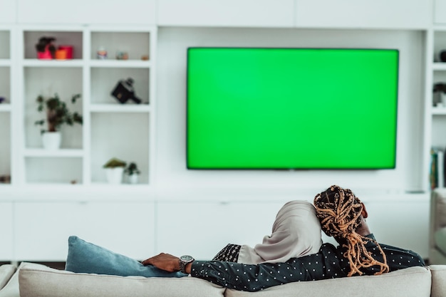 Afrykańska para siedzi na kanapie oglądając telewizję razem Chroma zielony ekran kobieta nosi ubrania islamskiego hidżabu. Zdjęcie wysokiej jakości