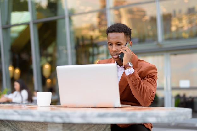 Afrykański biznesmen na zewnątrz w kawiarni przy użyciu laptopa podczas rozmowy przez telefon komórkowy