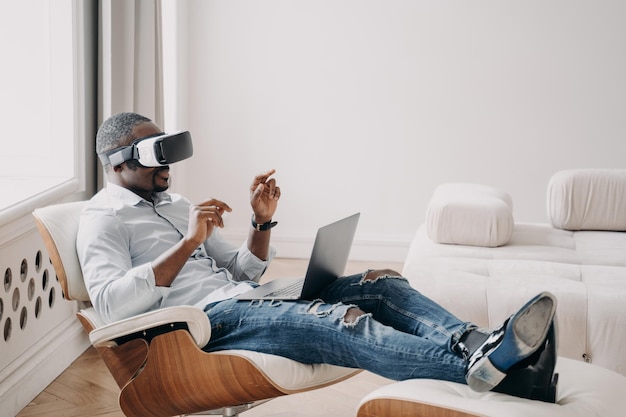 Afrykański biznesmen w okularach VR wchodzi w interakcję z wirtualną rzeczywistością siedzi w fotelu przy laptopie Zaawansowana technologia