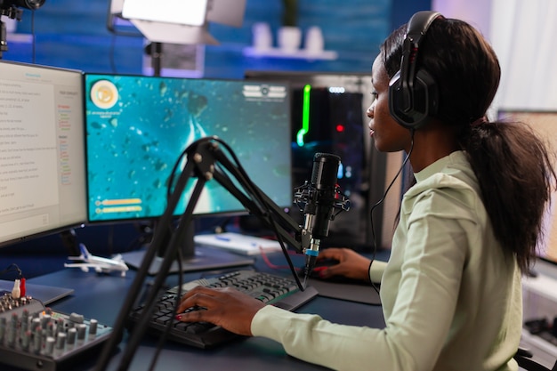 Afrykański gracz online e streamer próbuje wygrać kosmiczną strzelankę w ciemnym domu ze światłem rgb. Streamuj wirusowe gry wideo dla zabawy przy użyciu słuchawek i klawiatury podczas mistrzostw online.