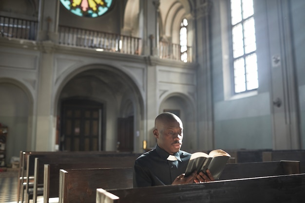 Afrykański ksiądz w kołnierzu siedzi na ławce w kościele i czyta Biblię