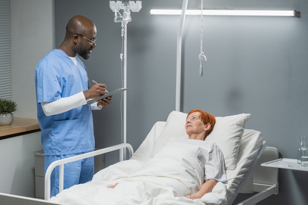 Afrykański lekarz-mężczyzna wypisujący receptę dla swojej starszej pacjentki, leżącej na łóżku na oddziale szpitalnym