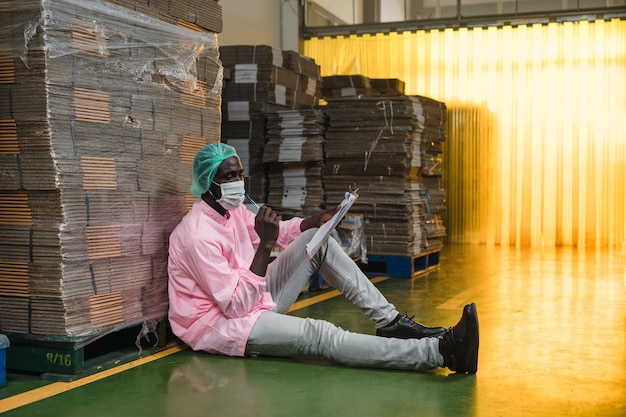 Afrykański męski kierownik kontroli zapasów w sterylnym mundurze siedzi i sprawdza kolejność opakowań kartonowych ułożonych w magazynie w zakładzie przetwórstwa napojów